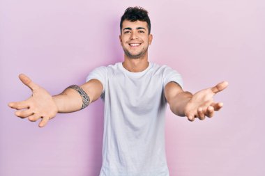 Latin asıllı genç bir adam, günlük beyaz tişört giyip kameraya bakıyor ve sarılmak için kollarını açmış gülümsüyor. Mutluluğu kucaklayan neşeli bir ifade.. 