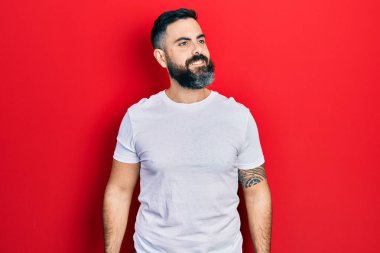 Latin kökenli genç bir adam günlük beyaz tişört giyiyor. Yan tarafa bakıyor, rahat bir görünüm takınıyor ve kendine güvenen bir gülümseme takınıyor.. 