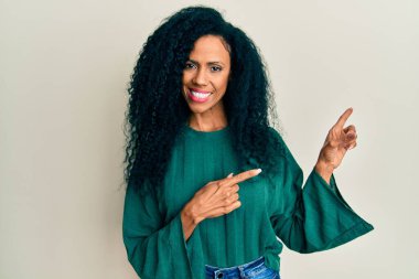 Orta yaşlı Afro-Amerikalı kadın günlük kıyafetler giyiyor gülümsüyor ve kameraya bakıyor iki eliyle ve parmaklarıyla yan tarafa bakıyor.. 