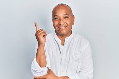 Orta yaşlı, sıradan kıyafetler giyen, yüzünde kocaman bir gülümseme olan, eli ve parmağı kameraya bakan Latin bir adam.. 