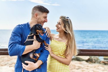 Kumsalda mutlu gülümseyen ve köpekle kucaklaşan beyaz bir çift..