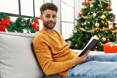 Genç Arap adam kitap okuyor. Evdeki Noel ağacının yanında, kanepede oturuyor..