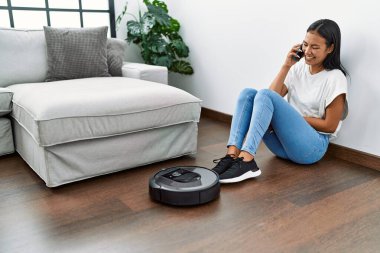 Akıllı telefondan konuşan genç Latin kadın evde akıllı robot süpürgesi kullanıyor.