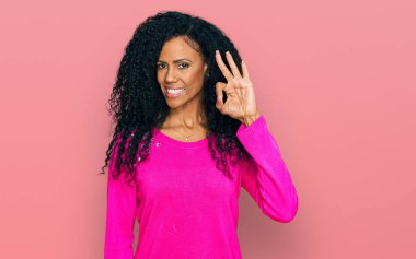Orta yaşlı Afro-Amerikalı kadın, günlük kıyafetler giyip gülümsüyor ve el ve parmaklarıyla 