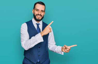 Sakallı genç adam iş yeleği giyiyor gülümsüyor ve kameraya bakıyor iki eliyle ve parmaklarıyla kenarı işaret ediyor. 