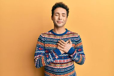 Kış süveteri giyen, elleri göğsünde, gözleri kapalı gülümseyen ve yüzünde minnet ifadesi olan genç yakışıklı bir adam. sağlık kavramı. 