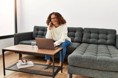 Orta yaşlı İspanyol kadın evde akıllı telefondan dizüstü bilgisayar kullanıyor.