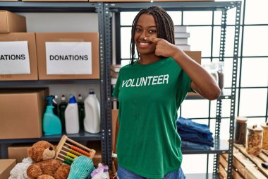 Bağışlarda gönüllü tişört giyen Afro-Amerikalı genç bir kadın el parmakları ve burunlarıyla gülümseyerek ayakta duruyor. güzellik konsepti 