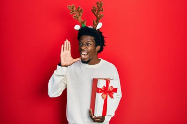 Geyik Noel şapkası takan genç Afrikalı Amerikalı adam elinde hediyeyle el ele bağırıyor ve el ele bağırıyor. iletişim konsepti. 