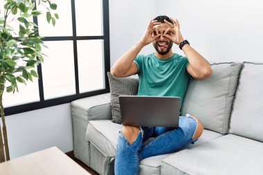 Sakallı genç yakışıklı adam evdeki kanepede oturup bilgisayar kullanıyor. Dürbün gibi jest yapıyor. Gözleri parmaklarından bakıyor. çılgın ifade. 