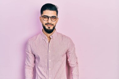 Sakallı, rahat gömlekli ve gözlüklü yakışıklı. Olumlu ve mutlu görünüyor. Kendine güvenen bir gülümsemeyle dişlerini gösteriyor. 