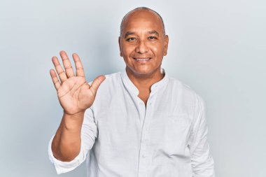 Orta yaşlı, gündelik kıyafetler giyen Latin bir adam beş numarayı gösterip işaret ederken kendinden emin ve mutlu bir şekilde gülümsüyor.. 