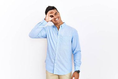 İş gömleği giyen genç İspanyol bir adam izole edilmiş arka planda duruyor. El hareketleriyle gülümsüyor, gözleriyle mutlu yüzle parmaklarının arasından bakıyor.. 