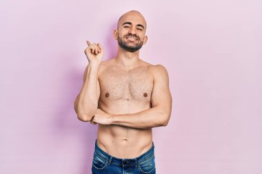 Tişörtsüz, yüzünde kocaman bir gülümsemeyle genç ve kel bir adam, eli ve parmağı kameraya bakıyor.. 