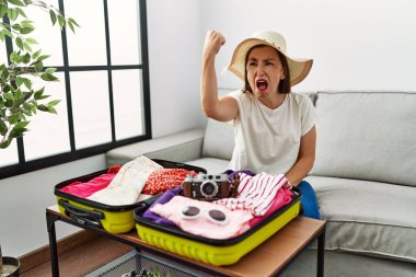 Güzel, orta yaşlı İspanyol bir kadın, kızgın ve kızgın bir şekilde bavuluna yazlık giysiler yerleştiriyor ve öfkeyle bağırırken yumruğunu kaldırıyor. öfke ve agresif konsept. 