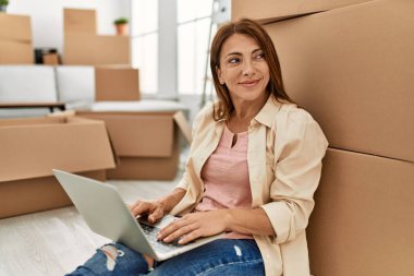 Orta yaşlı, beyaz bir kadın yeni evinde dizüstü bilgisayar kullanıyor..