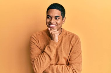 Elleri çenesinde gündelik kazak giyen, soru soran, düşünceli, genç ve yakışıklı İspanyol bir adam. Düşünceli bir yüzle gülümsüyor. şüphe kavramı. 