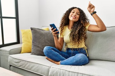 Güzel İspanyol kadın evinde kanepede oturmuş akıllı telefon kullanıyor. Gülümsüyor, şaşırıyor, parmaklarıyla işaret ediyor ve kollarını kaldırıyor.. 