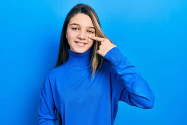 Balıkçı yaka kazak giyen genç esmer kız, parmaklarıyla yüzünü ve burnunu göstererek neşeli bir şekilde gülümsüyor. güzellik konsepti 