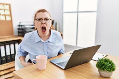 Ofiste çalışan genç, kızıl saçlı bir kadın bilgisayar laptopu kullanıyor kızgın ve öfkeli çığlıklar atıyor öfkeyle bağırıyor. öfke ve agresif konsept. 