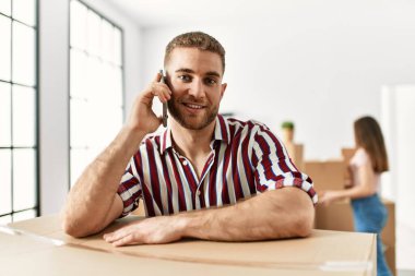 Genç İspanyol adam akıllı telefondan konuşuyor. Yeni evde karton üzerine yaslanıyor..
