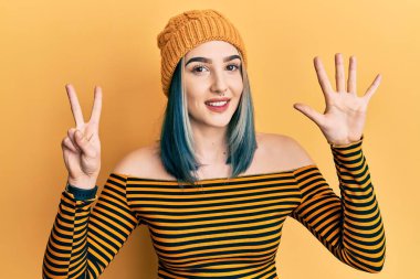 Genç modern kız yün şapka takıyor ve yedinci parmaklarını gösterip işaret ederken kendinden emin ve mutlu gülümsüyor.. 