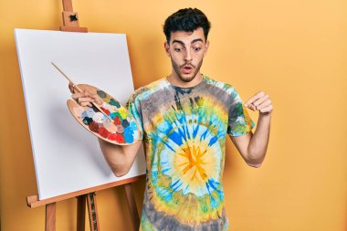 İspanyol asıllı genç adam palet üstüne palet çizerek ayakta duruyor parmaklarını göstererek reklamını yapıyor, şaşırmış yüzünü ve ağzını açıyor. 