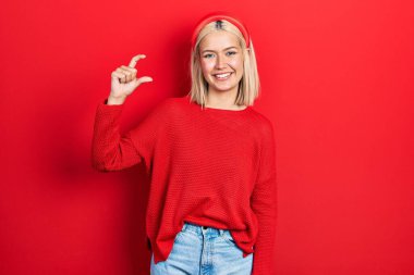 Güzel sarışın kadın, günlük kırmızı süveter giyiyor gülümsüyor ve kendine güler yüzlü el hareketleriyle küçük beden işareti yapıyor parmaklarıyla bakıyor ve kameraya bakıyor. ölçüm kavramı. 