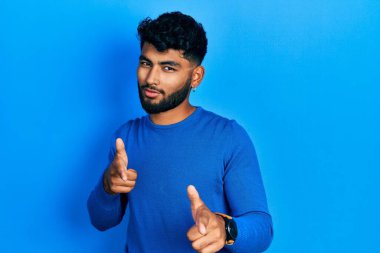 Sıradan mavi süveter giyen sakallı bir Arap mutlu ve komik yüzlü parmaklarını kameraya doğrultuyor. iyi enerji ve titreşimler. 