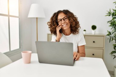 Orta yaşlı İspanyol kadın evde akıllı telefondan dizüstü bilgisayar kullanıyor.