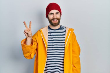 Sakallı, sarı yağmurluk giyen, kameraya gülümseyen ve parmaklarıyla zafer işareti yapan beyaz bir adam. İki numara.. 