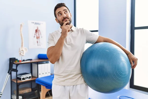 Przystojny Latynos Trzymający Pilates Ball Klinice Rehabilitacyjnej Poważna Twarz Myślący — Zdjęcie stockowe