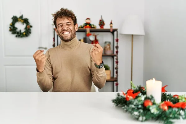 年轻英俊的男人 留着胡子 坐在桌子上 圣诞装饰品让他很开心 很兴奋 他举起双臂做了获胜的手势 微笑着 尖叫着要成功 庆祝概念 — 图库照片