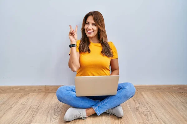 西班牙妇女用笔记本电脑坐在家里的地板上 笑容满面地看着摄像机用手指做胜利的标志 第二点 — 图库照片