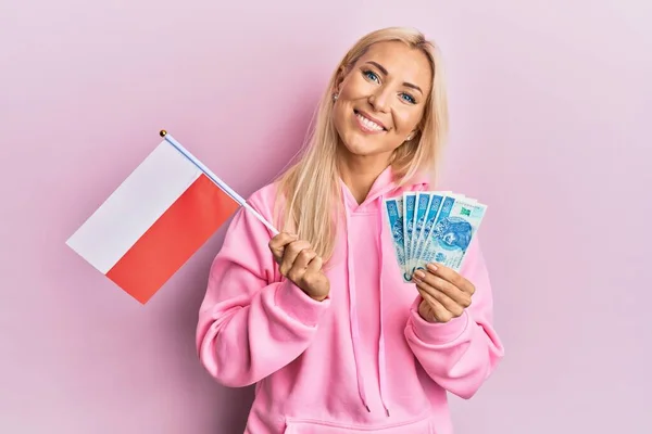 年轻的金发女子举着波兰国旗和兹罗提钞票 脸上挂着快乐而沉着的微笑 显示牙齿 — 图库照片