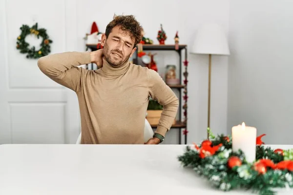 年轻英俊的男人 留着胡子 坐在桌子上 戴着圣诞装饰品 脖子酸痛 用手触摸脖子 肌肉疼痛 — 图库照片