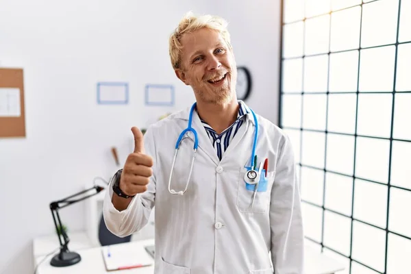 年轻的金发男子穿着医生制服和听诊器在诊所做着快乐的大拇指举手表决 赞成的表情看着相机显示的成功 — 图库照片