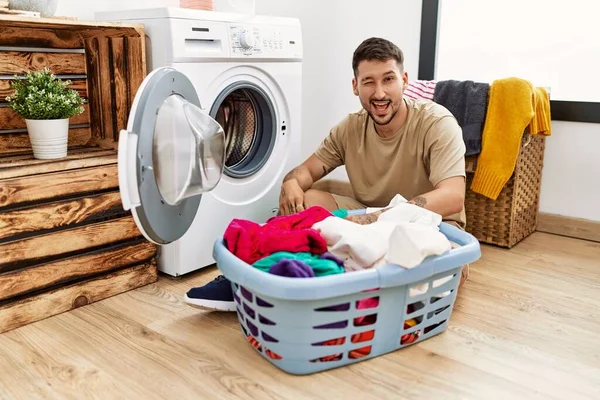 年轻英俊的男子把脏衣服放进洗衣机里眨眼看着摄像机 脸上带着性感的表情 笑容满面 — 图库照片