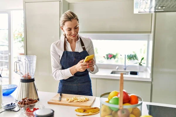 年轻的金发女人在厨房用智能手机切香蕉 — 图库照片