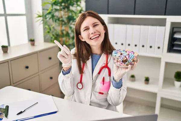 お菓子を持っている若い医師の女性は側に手と指で笑顔幸せなポインティング — ストック写真