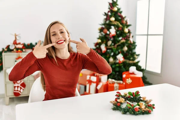 Kavkazská Mladá Blondýnka Sedící Stole Vánočního Stromku Vesele Usmívající Ukazujíc — Stock fotografie
