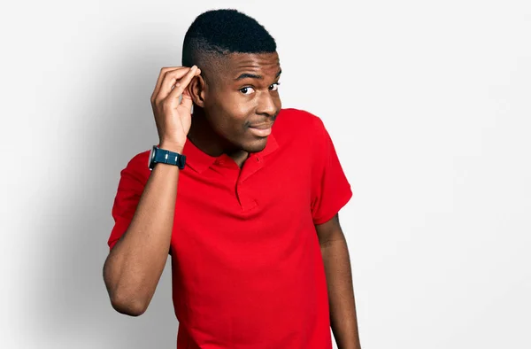 噂やゴシップに耳を傾ける耳の上に手で笑みを浮かべてカジュアルな赤いTシャツを着て若いアフリカ系アメリカ人の男性 聴覚障害の概念 — ストック写真