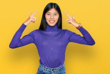 Güzel, genç Asyalı kadın günlük kıyafetler giyip gülümsüyor iki eliyle başını işaret ediyor, harika bir fikir ya da düşünce, iyi bir hafıza. 