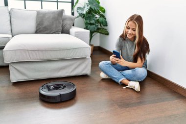 Evde yerde oturan akıllı telefonuyla robot elektrik süpürgesini kontrol eden beyaz bir kız..