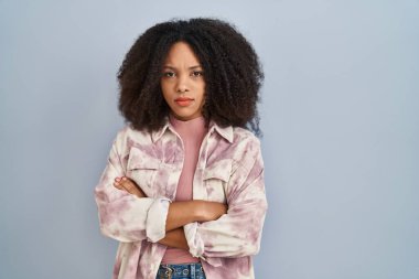 Mavi arka planda duran genç Afrikalı Amerikalı kadın şüpheci ve gergin, yüzünde çapraz kollarla onaylamayan ifade. negatif kişi. 