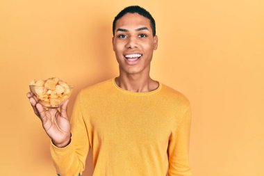 Genç Afro-Amerikalı adam elinde patates cipsi tutuyor. Olumlu ve mutlu görünüyor. Kendine güvenen bir gülümsemeyle dişlerini gösteriyor. 