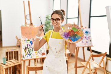 Resim stüdyosunda elinde fırça ve palet tutarak gülümseyen sevimli kız.
