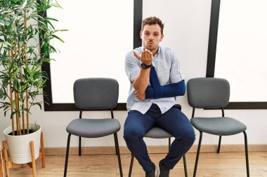 Yakışıklı genç adam, doktor bekleme odasında kolundan yaralanmış bir şekilde oturuyor kameraya bakıyor ve havayla öpüşüyor. Güzel ve seksi. aşk ifadesi. 