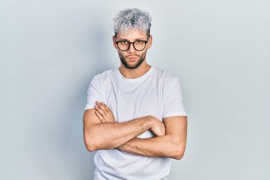 Modern boyalı saçlı, beyaz tişörtlü, gözlüklü, şüpheci ve gergin, yüzünde çapraz kollu tasvip etmeyen bir ifade olan genç bir İspanyol. negatif kişi. 