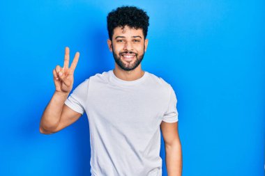Sakallı, beyaz tişörtlü genç Arap adam kameraya bakıp zafer işareti yapıyor. İki numara.. 
